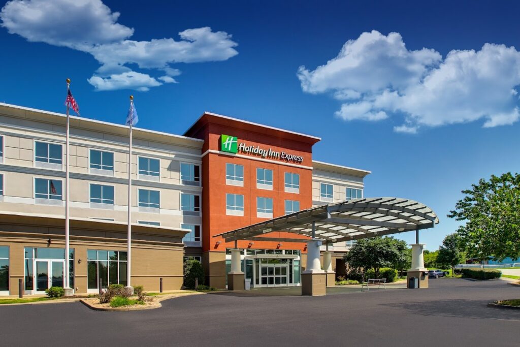Exterior view of a Holiday Inn Express hotel with a covered entrance, flagpoles out front, and a blue sky with scattered clouds.